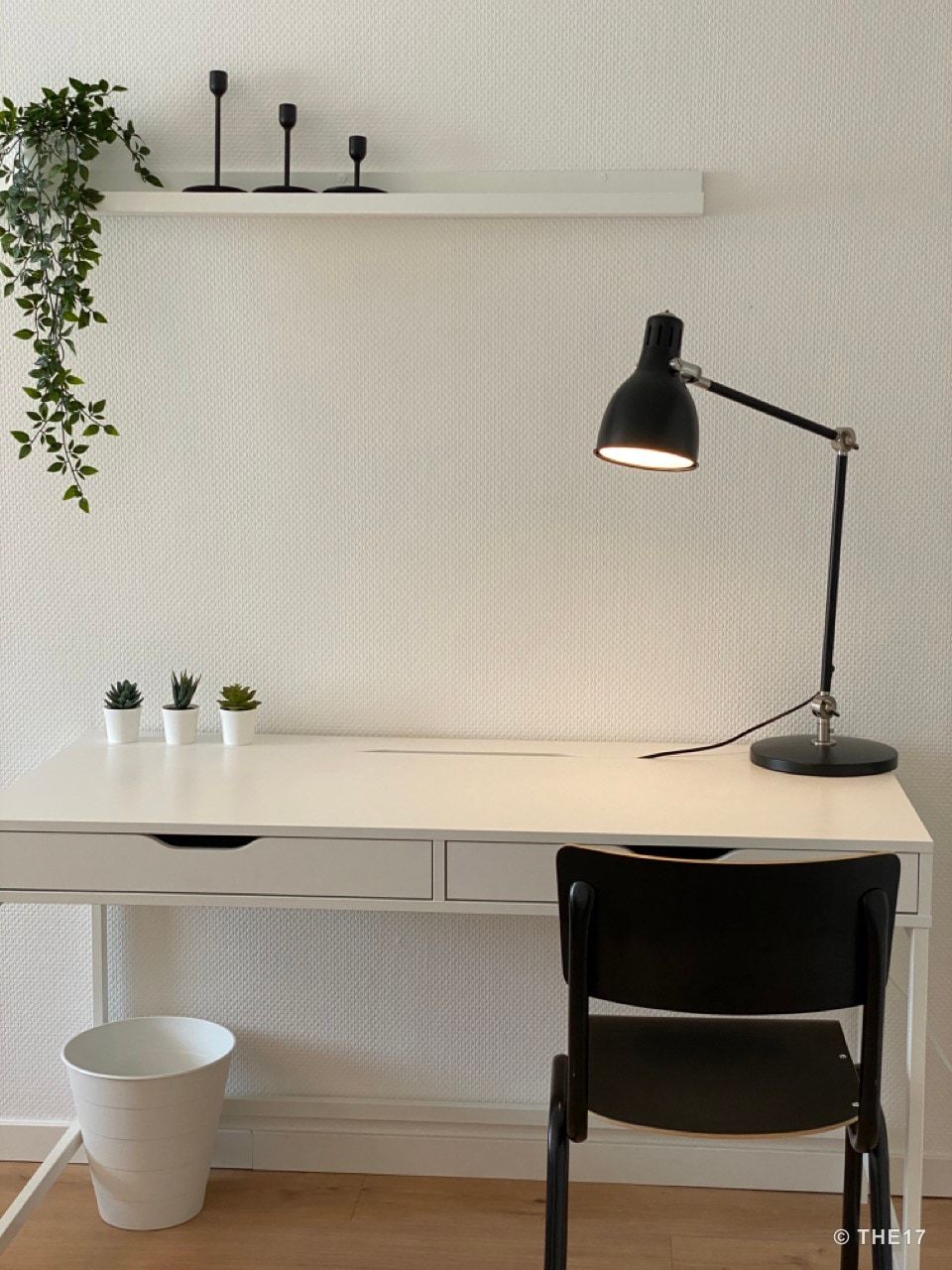Moderner Arbeitsplatz mit weißem Schreibtisch, schwarzem Stuhl, Schreibtischlampe und kleinen Pflanzen vor weißer Wand.